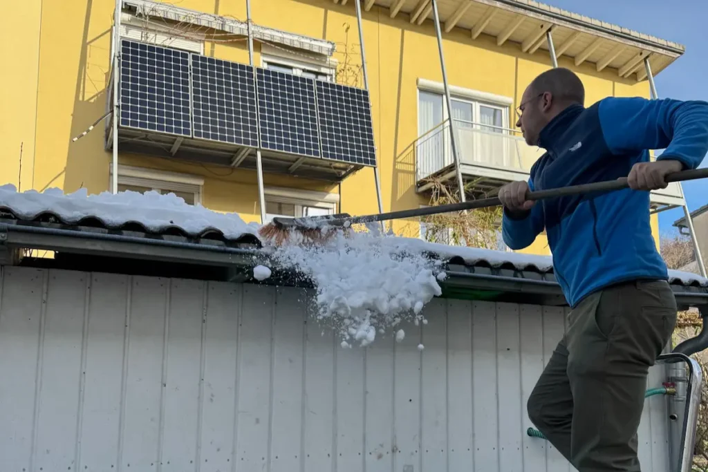 Mann steht auf Leiter und räumt mit Besen Schnee vom Dach.