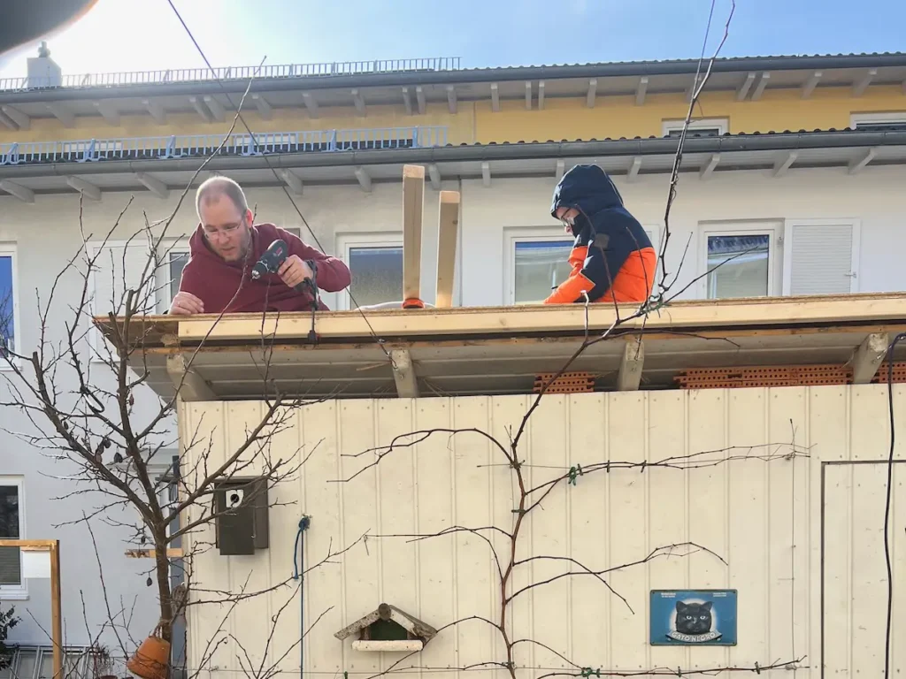 Zwei Personen sitzen auf dem Dach eines Gartenhauses und montieren eine Holzleiste.